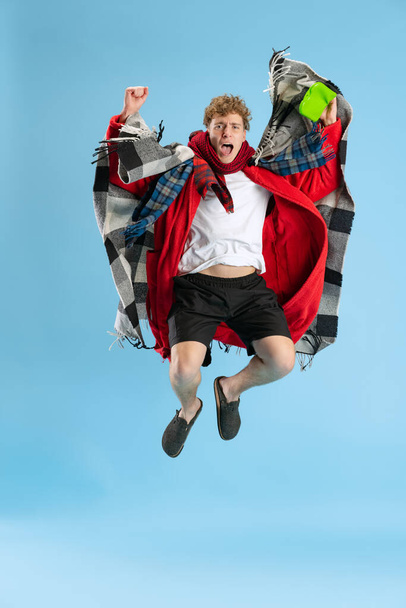Full-length portrait of sick young boy, wearing plaid, robe and scarf joyfully jumping over blue background. Recovering from cold and flu, allergy - Foto, Imagem