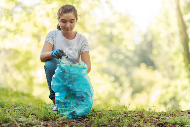 Young volunteer cleaning up the forest, she is collecting trash, environmental protection concept - Фото, изображение