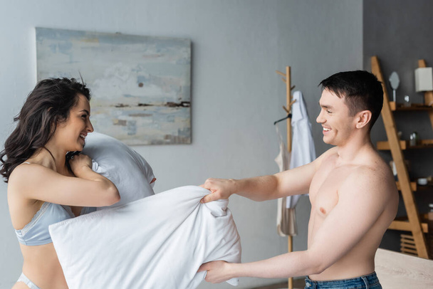 side view of joyful and sexy couple having pillow fight in bedroom  - Foto, Imagem