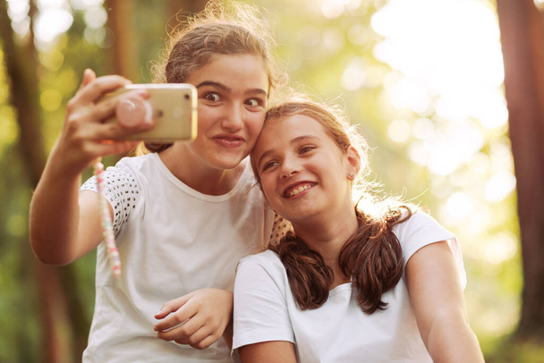 Ormanda birlikte poz veren tatlı genç kızlar akıllı telefonlarıyla selfie çekiyorlar. - Fotoğraf, Görsel