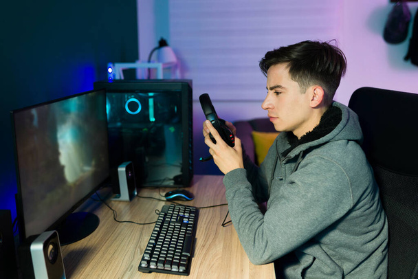彼のコンピュータ上でオンラインビデオゲームをプレイし始めるために彼のヘッドフォンに置くハンサムな男性ゲーマー - 写真・画像