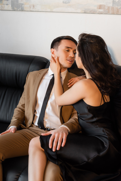 brunette woman in black slip dress touching face of man on sofa  - Fotó, kép
