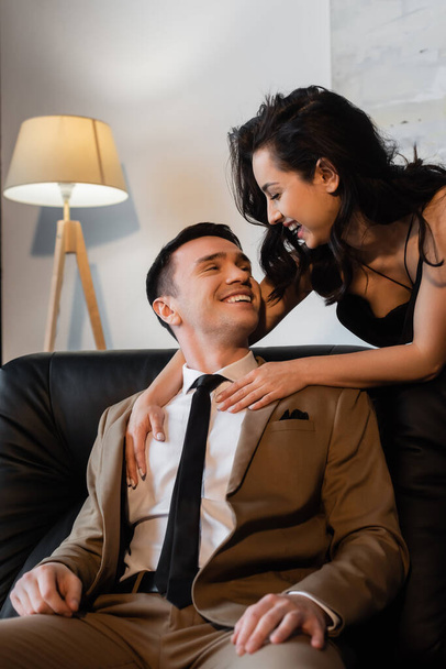 szczęśliwy brunetka kobieta przytulanie uśmiechnięty mężczyzna na kanapie  - Zdjęcie, obraz