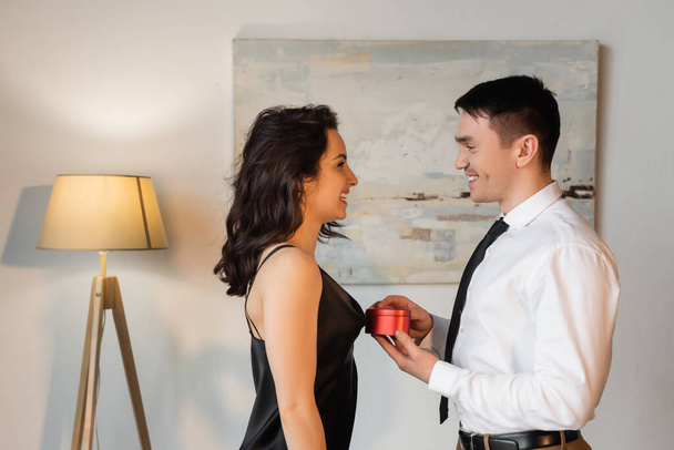 boční pohled na muže drží srdce ve tvaru dárkové krabice v blízkosti šťastný přítelkyně v černé kombiné šaty  - Fotografie, Obrázek