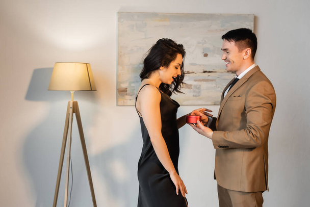 Seitenansicht des glücklichen Mannes mit herzförmiger Geschenkschachtel in der Nähe der erstaunten Freundin im schwarzen Slip-Kleid  - Foto, Bild