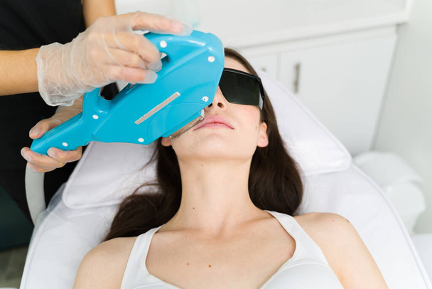 Ελκυστική γυναίκα πελάτης να πάρει μια αισθητική διαδικασία για να αφαιρέσετε τα μαλλιά στο πάνω χείλος της με έντονο παλλόμενο φως - Φωτογραφία, εικόνα