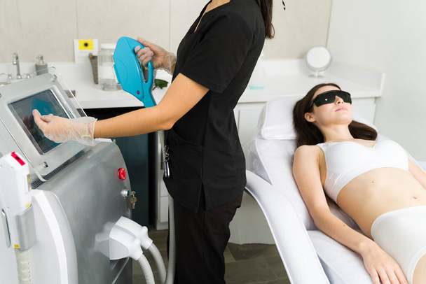 esteticista trabajando en cosmetología preparándose para hacer un tratamiento de depilación láser para una cliente femenina en el spa - Foto, imagen