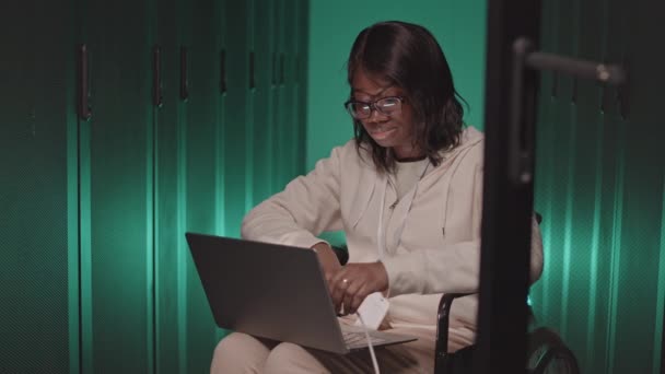 Moyenne longue de jeune femme afro-américaine en fauteuil roulant, tenant l'ordinateur portable sur les genoux dans le centre de données vert-éclairé, branchant le câble dans l'armoire de serveur - Séquence, vidéo
