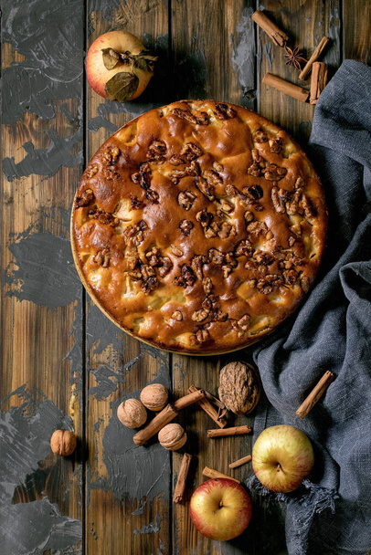 Sweet homemade apple and walnuts cake Charlotte on plate with fresh gardening apples, cinnamons and nuts around over dark wooden plank background. Autumnal home baking. Flat lay, copy space - Φωτογραφία, εικόνα