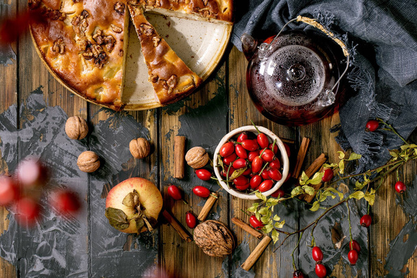 Homemade apple and walnuts cake Charlotte on plate with fresh gardening apples, nuts, rose hip tea around over dark wooden plank background. Autumnal home baking. Flat lay - Фото, зображення