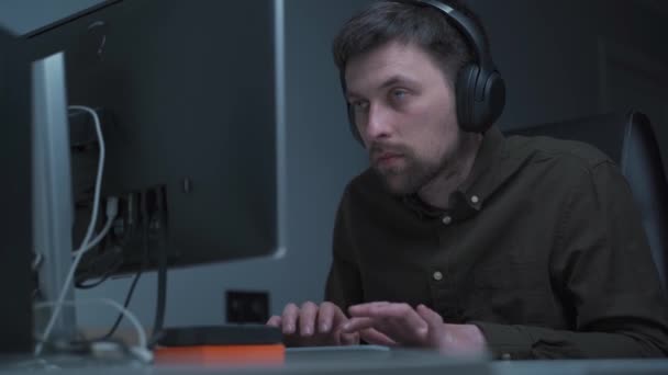 Application developer in wireless headphones working. sitting at computer workplace in home office. IT guy focused and tiredly looking at monitor while programming. Overtime, deadlines are on fire - Footage, Video
