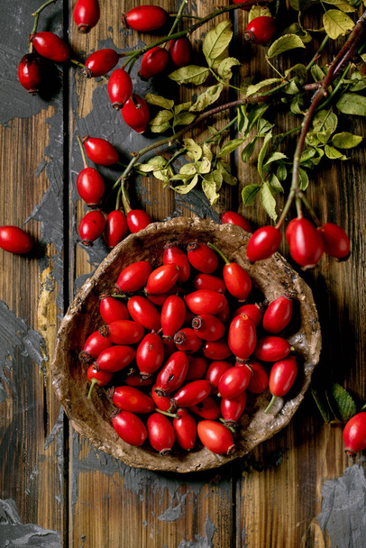 Rose hip berries on ceramic plate with branch and leaves on old wooden plank background. Autumn reserves of vitamins for the winter. Flat lay, close up - Photo, Image