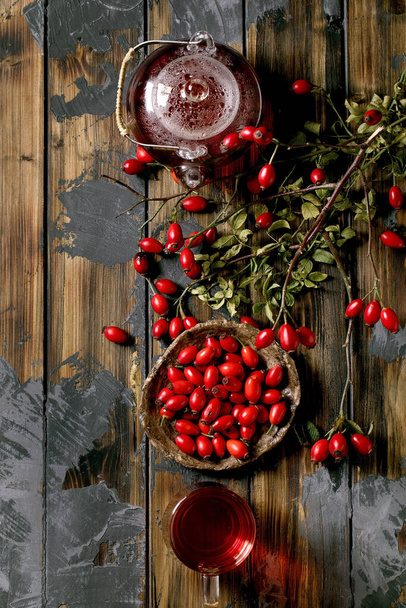 Rose hip berries herbal tea in glass teapot and cup standing on old wooden plank background with wild autumn berries around. Winter hot beverage. Flat lay - Foto, immagini