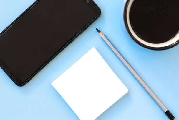 Flat lay com um smartphone, nota de papel branco, uma xícara de café e lápis sobre fundo azul claro. Preparem-se, espaço de cópia. - Foto, Imagem