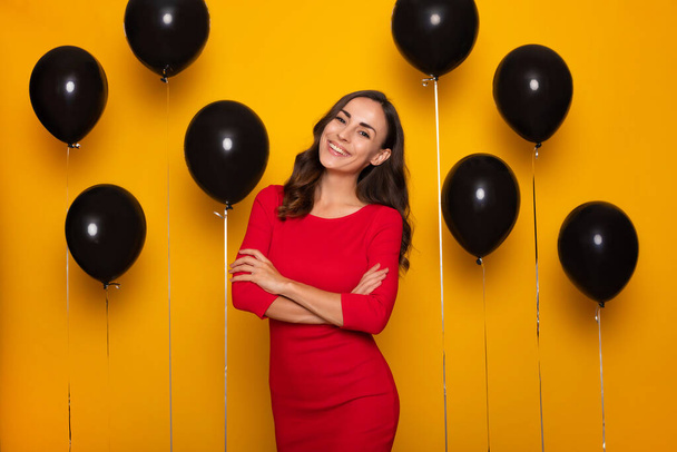 Close up photo of an attractive smiling confident brunette woman in red dress on black helium balloons background in sales day - Фото, изображение
