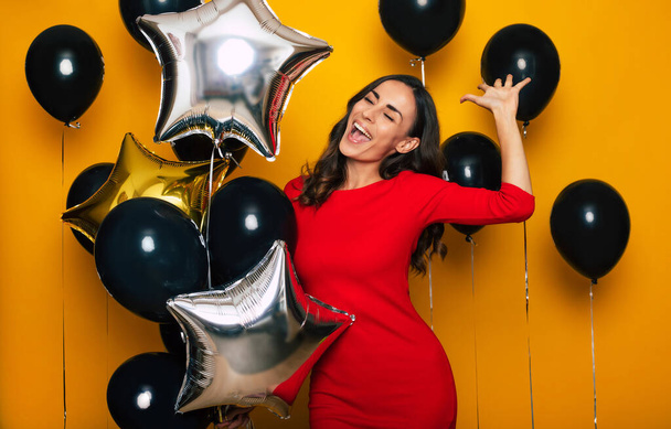 Close up photo of an excited cute fashionable young woman in a trendy red dress while she is having fun and posing with the many balloons in black Friday on yellow background - Photo, Image