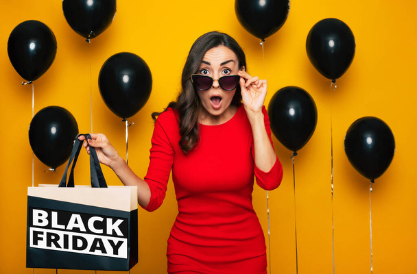 Fashionable happy gorgeous lady with a shopping bag in hand is posing in a good excited mood in the background of many helium balloons in black Friday - Фото, изображение
