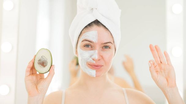 A woman with a towel on her hair and in a clay face mask holds an avocado. Taking care of beauty at home - Foto, Bild