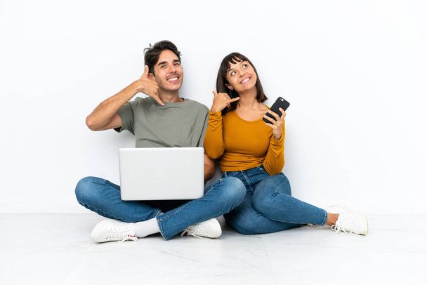 Young couple with a laptop and mobile sitting on the floor making phone gesture. Call me back sign - Photo, Image