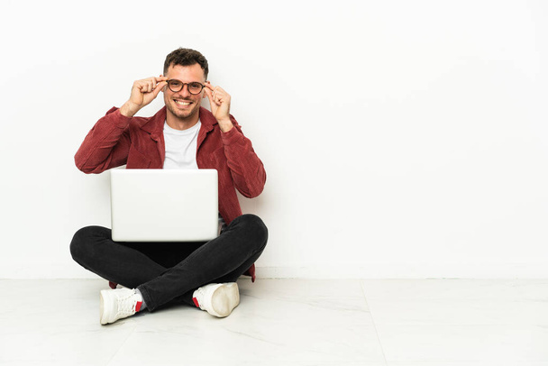 Giovane uomo caucasico bello sit-in sul pavimento con computer portatile con occhiali e sorpreso - Foto, immagini
