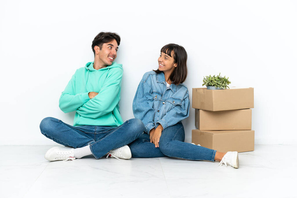 Молода пара робить крок, підбираючи коробку, повну речей, що сидять на підлозі, ізольовані на білому тлі, дивлячись через плече з посмішкою
 - Фото, зображення