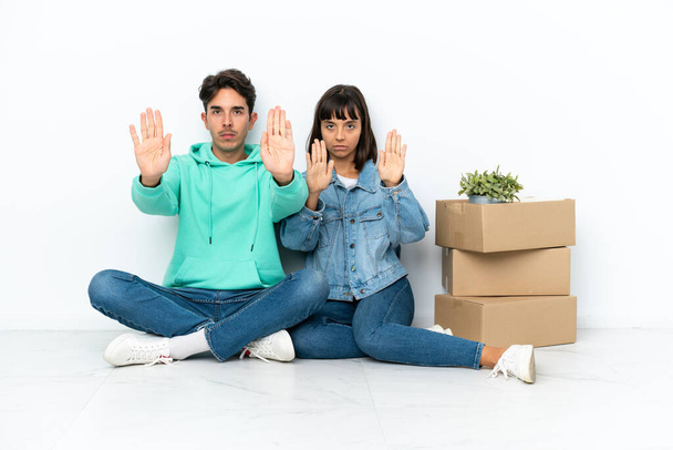 Молодая пара делает шаг, поднимая коробку, полную вещей, сидя на полу изолированы на белом фоне делает остановку жест для разочарован мнением - Фото, изображение