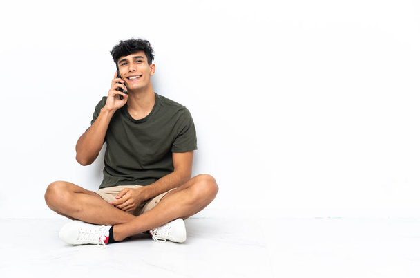 Mladý argentinský muž sedí na podlaze a vede rozhovor s mobilním telefonem - Fotografie, Obrázek