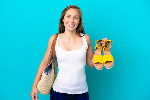 Young caucasian woman holding summer sandals isolated on blue background smiling a lot - Photo, Image