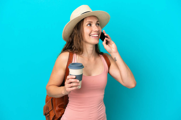 Junge kaukasische Frau in den Sommerferien isoliert auf blauem Hintergrund mit Kaffee zum Mitnehmen und einem Handy - Foto, Bild