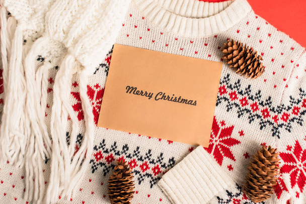 top view of greeting card with merry christmas lettering near pine cones on knitted sweater  - Zdjęcie, obraz
