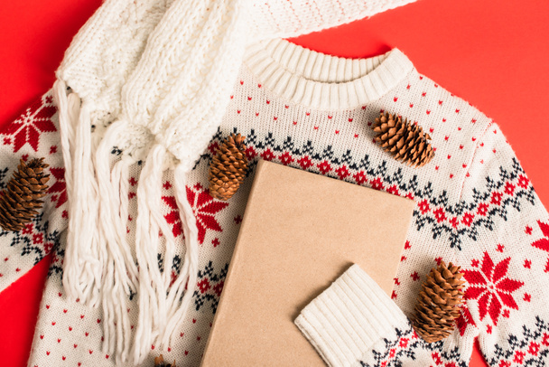 top view of book near pine cones on knitted sweater and red background - Fotó, kép