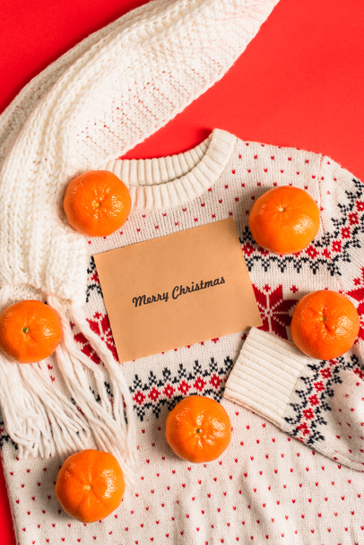 top view of greeting card with merry christmas lettering near tangerines on knitted sweater isolated on red - Photo, Image