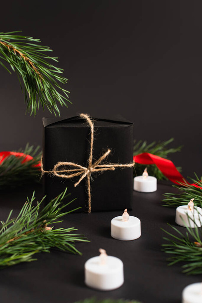 caja de regalo envuelta cerca de ramas de abeto y velas con cinta roja borrosa sobre fondo negro - Foto, imagen