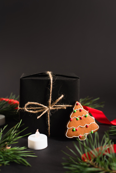 завернутый подарок рядом с еловыми ветвями, пряничное печенье и свеча на черном  - Фото, изображение