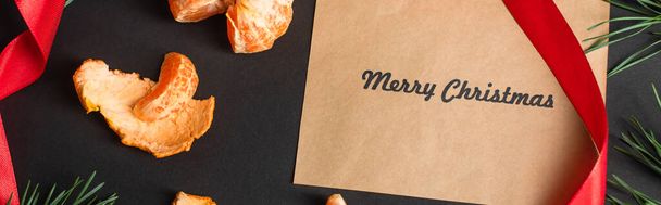 top view of craft paper with merry christmas lettering near peeled tangerines on black, banner - Фото, изображение