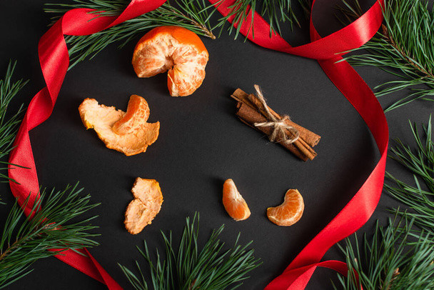 top view of peeled tangerines and cinnamon sticks near red ribbon and fir branches on black - Photo, Image