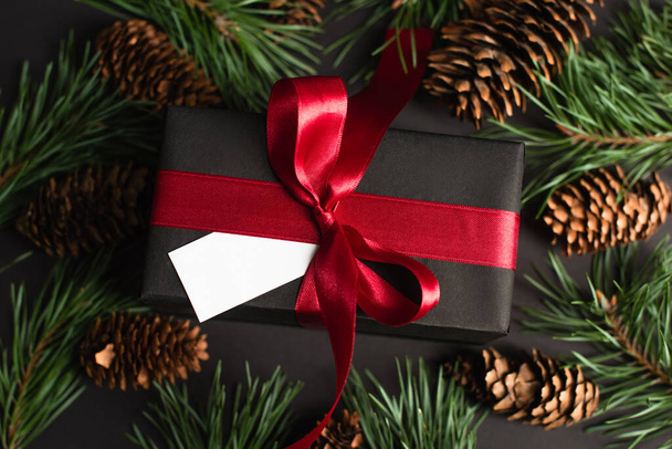 vista superior de la caja de regalo envuelta con cinta roja y tarjeta en blanco cerca de ramas de abeto y conos de pino en negro - Foto, Imagen