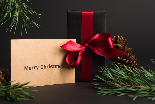 craft χαρτί με χαρούμενα χριστουγεννιάτικα γράμματα κοντά κουτί δώρου και κλαδιά ελάτης σε μαύρο - Φωτογραφία, εικόνα