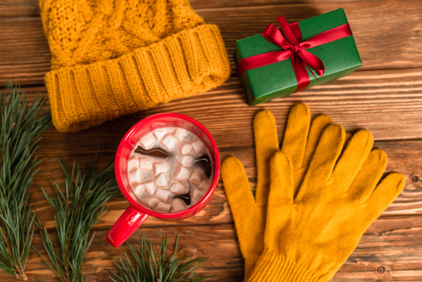 top view of yellow knitted hat and gloves near gift box, cup of cocoa with marshmallows and branches on wooden surface - Valokuva, kuva