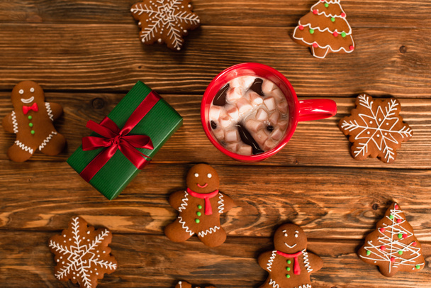top view of cup of cocoa with mályvacukor near gift box and mingerbread cookies on wooden surface - Fotó, kép