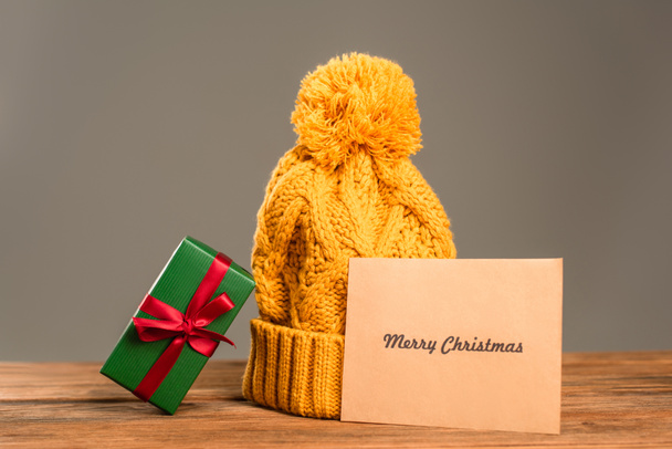 Grußkarte mit frohen Weihnachten Schriftzug in der Nähe Strickmütze und eingewickeltes grünes Geschenk isoliert auf grau - Foto, Bild