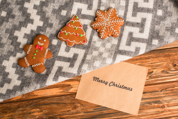 κορυφαία άποψη των μπισκότων μελόψωμο σε γκρι κουβέρτα με στολίδι κοντά ευχετήρια κάρτα με χαρούμενα γράμματα Χριστούγεννα σε ξύλινη επιφάνεια  - Φωτογραφία, εικόνα