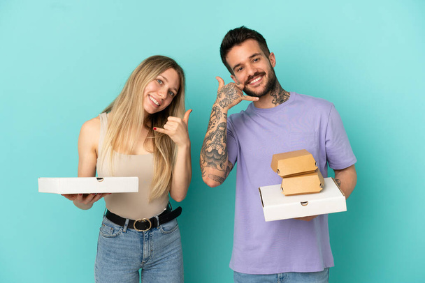 Пара держащих пиццу и бургеры на изолированном синем фоне, делая телефонный жест. Перезвони мне - Фото, изображение