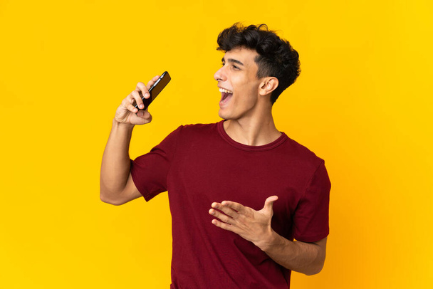 Young Argentinian man isolated on yellow background using mobile phone and singing - Zdjęcie, obraz