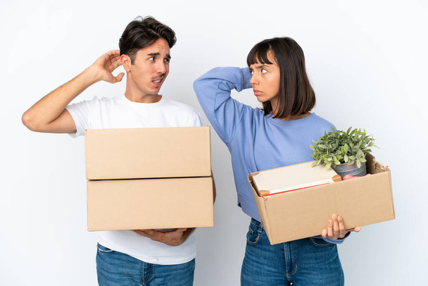 Young couple making a move while picking up a box full of things isolated on white background having doubts while scratching head - Photo, Image