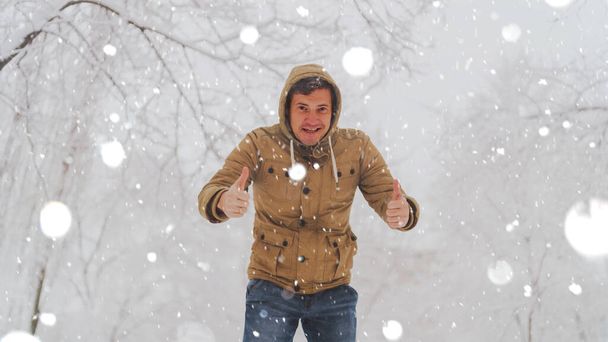 Handsome man standing and showing thumbs up in park in winter season. Adult young male wearing beige hooded jacket showing gesture of approval in snowy day. - Valokuva, kuva