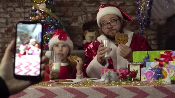 Ένας άνδρας ντυμένος Άγιος Βασίλης και ένα μικρό κορίτσι πίνουν γάλα και cookies.Posing για smartphone. - Πλάνα, βίντεο