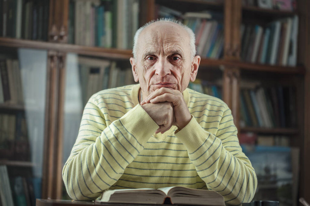 Senior male author sitting at table with book and holds hands under head. - Foto, Imagem