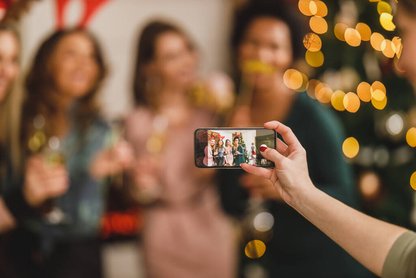 一緒に家で新年を祝うように、楽しい時間を過ごし、スマートフォンで思い出の写真を撮る陽気な多国籍女性の友人のグループ。スマートフォンのクローズアップ. - 写真・画像