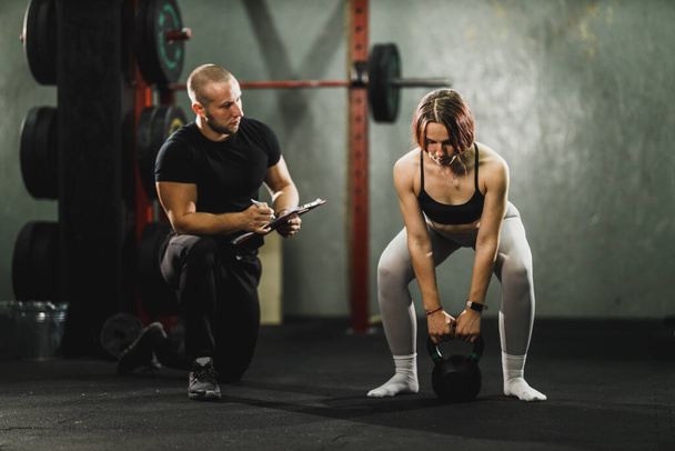 Izmos fiatal nő edz az edzővel az edzőteremben. Felpumpálja a muszkulumát kettlebell-lel.. - Fotó, kép
